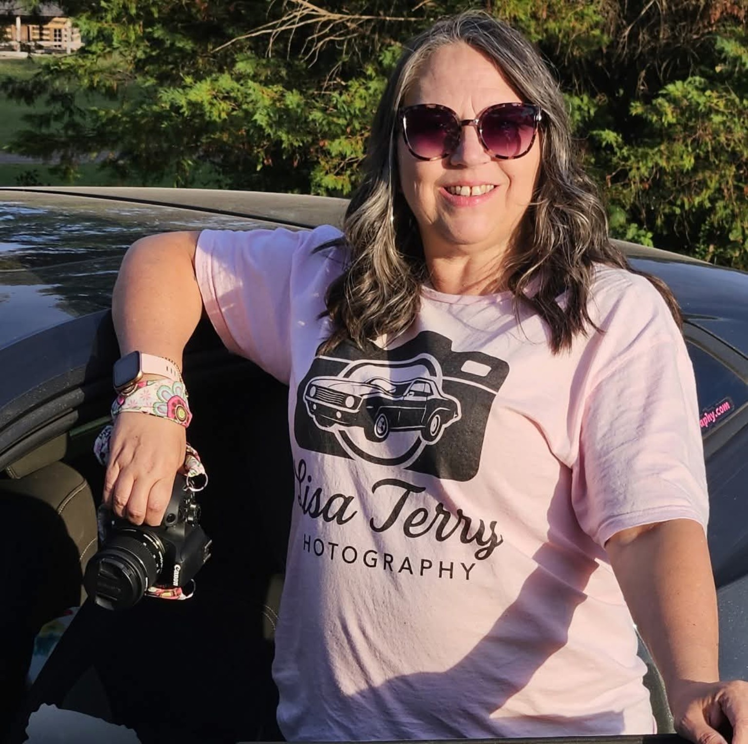 Lisa Terry holding her Canon camera beside a Camaro and wearing her Lisa Terry Photography shirt